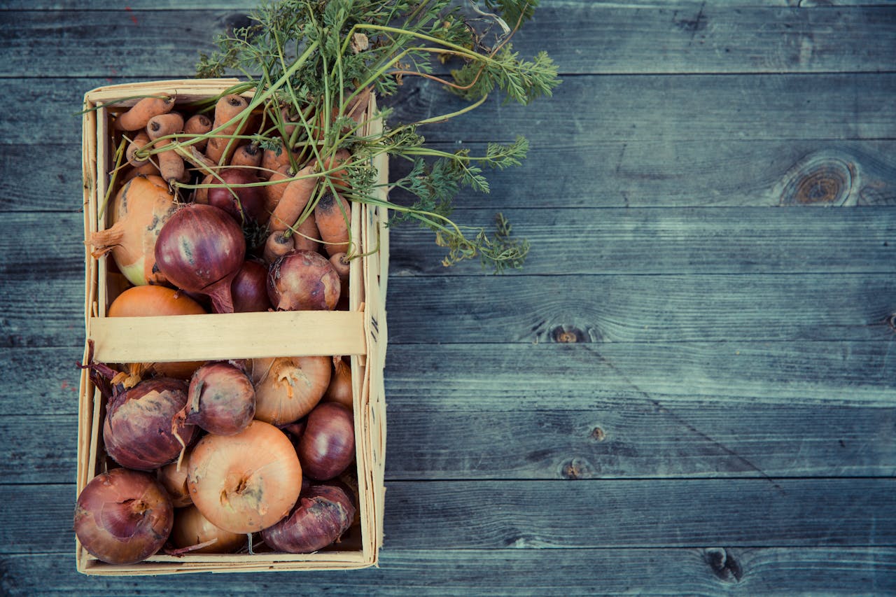 Basket of fresh onions and carrots on a rustic wooden surface, showcasing a farm-to-table harvest.