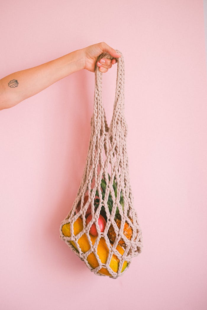 Hand holding a mesh bag filled with colorful fruits against a pink background.