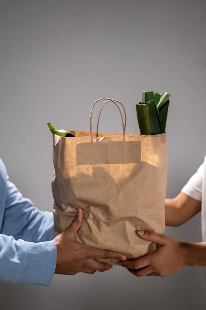 Close-up of hands exchanging a brown paper grocery bag filled with vegetables.